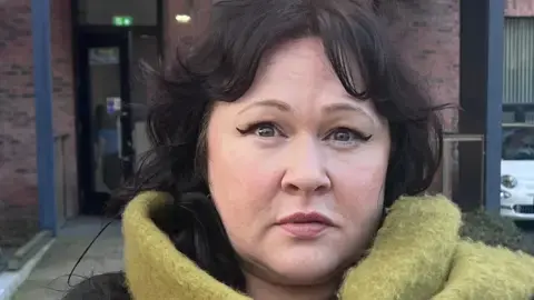 سارا موریسون ایستاده در خارج از ورودی یک ساختمان دولتی از آجرهای قرمز. موهای تیره‌اش که تا شانه می‌رسد، به‌صورت میانی تقسیم شده و حالت چهره‌اش خنثی است. او پیراهن بلند آستین سیاه به تن داشته و شال پشمی سبز رنگ دور گردن و بر شانه‌اش پیچیده است.