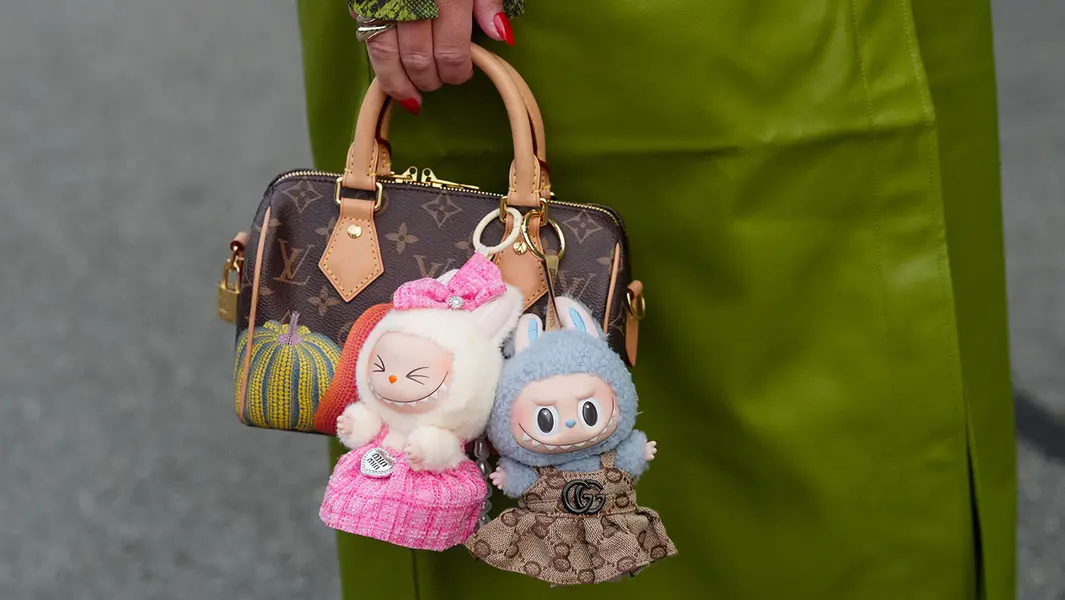 یک مهمان کت چرمی روشن به رنگ زیتونی پوشیده که مچسرپنبهاش طرح مار سبز دارد و از آستین بیرون میآید، یک حلقه نقرهای رنگ و مانیکور براق قرمز نوکتیز دارد؛ در عین حال کیف مینی با دسته بالای بوم منوگرام لوئیویتون به رنگ قهوهای، با دستگیرههای موچیده از چرم واهتا طوسی، برچسبهای چرمی، زیپ طلایی و قفل طلا، و طرح کدوی زرد‑نارنجی حمل میشود؛ دو نماد عروسکی لابوبو به کیف وصل شدهاند — یکی یک عروسک کرمی با لباس نخی صورتی و کمان هماهنگ، دیگری عروسکی خاکستری‑آبی با گوشهای خرگوش در لباس قهوهای منوگرام گوچی GG که با نشان دو G سیاه و جزئیات طلایی تکمیل شده است. صحنهای بیرون از Rotate، در هفتهٔ مد کپنهاگ، در تاریخ ۷ آگوست ۲۰۲۵ در کپنهاگ، دانمارک.