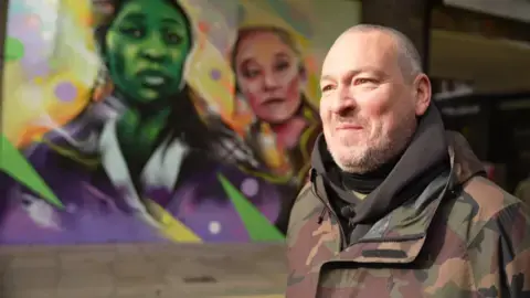 آقای سنز در مقابل دیوارنگارهٔ خود ایستاده است که شخصیت‌های اصلی ویکد را نشان می‌دهد