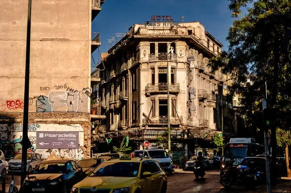 نمایی از خیابان که یک ساختمان فرسوده هتل را با تابلوی قرمز «Hotel Sans Rival» در بالای آن نشان می‌دهد. خودروها و یک موتورسیکلت از کنار دیواری پوشیده از گرافیتی عبور می‌کنند.